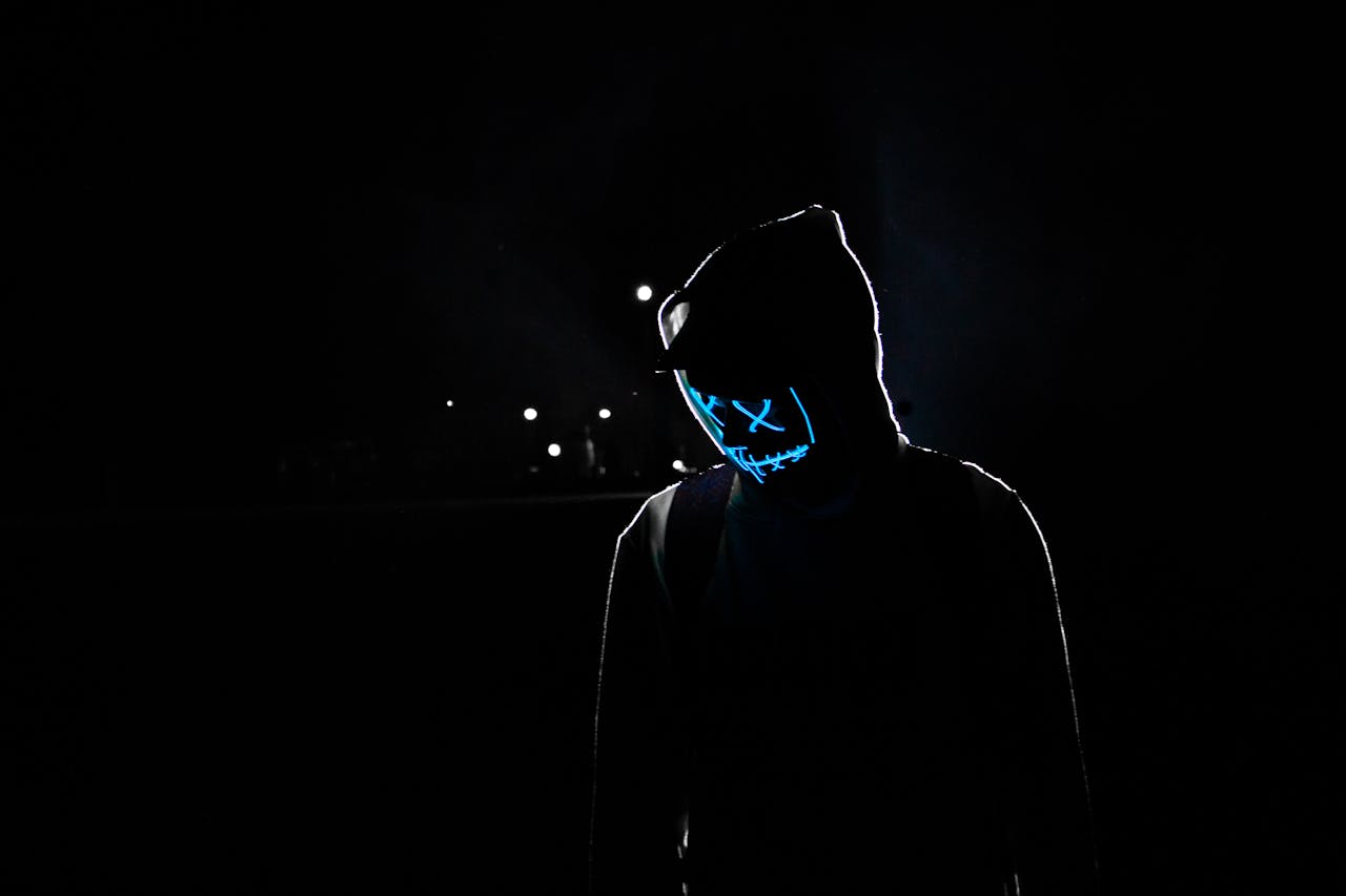 Silhouette of a person in a hoodie wearing a glowing LED mask against a dark background.